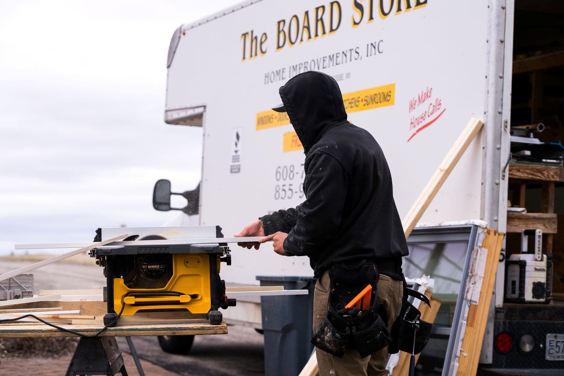 A Board Store worker cuts material with a table saw in front of a Board Store truck.