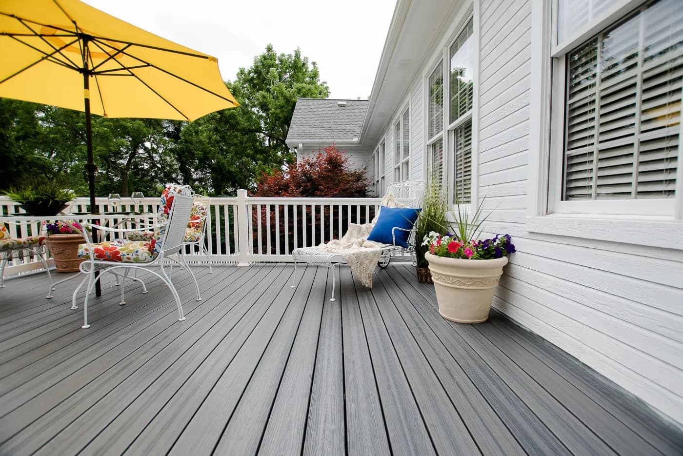 Tan house with a brown deck on the first floor with a green lawn and woods