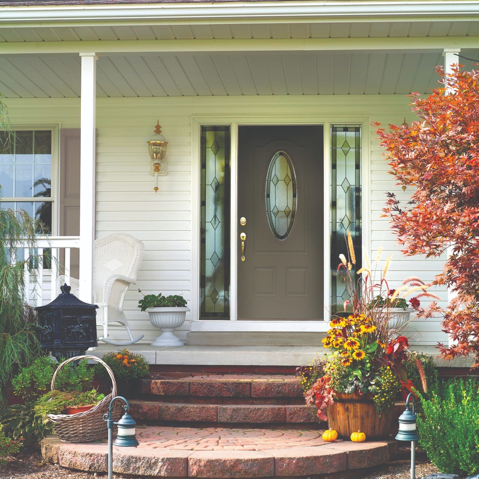 Front porch of cream-colored house with dark front door, decorative sidelights, white columns, white wicker chair, fall foliage, mums, pumpkins, and autumn landscaping