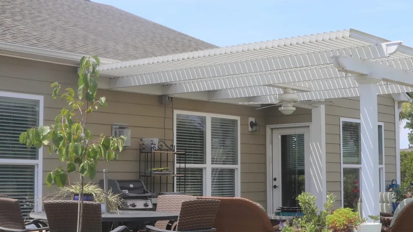 A white pergola attached to a home with brown siding and white trim doors and windows