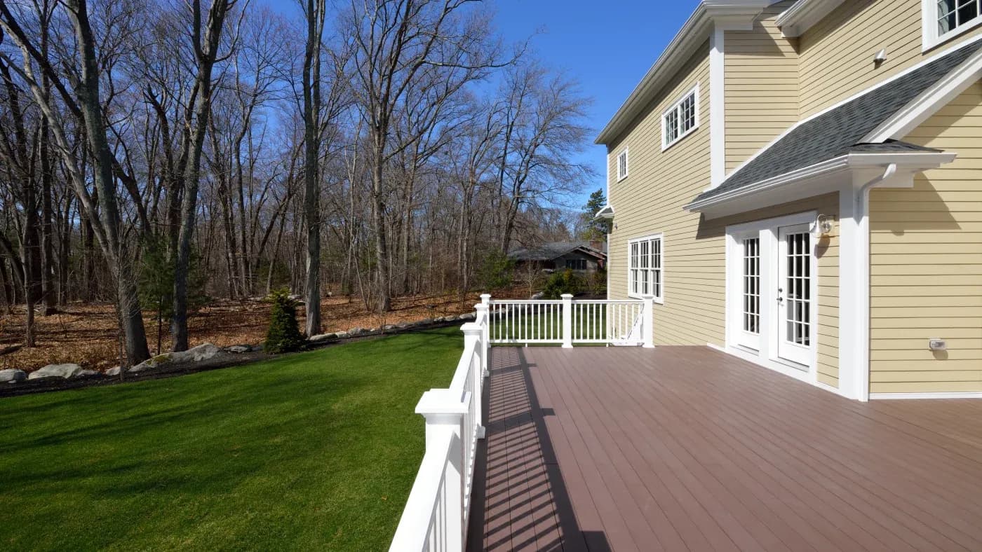 Tan house with a brown deck on the first floor with a green lawn and woods